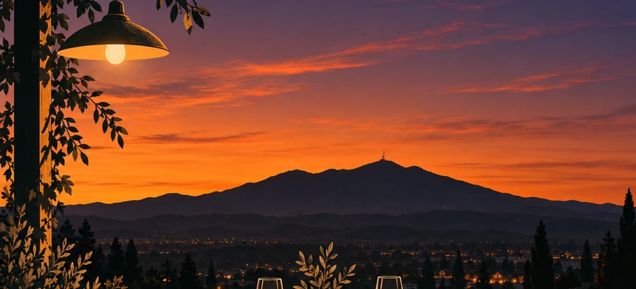 Illustration: a lamp and two wine glasses on a terrace at sunset, a mountain and town lights beyond.