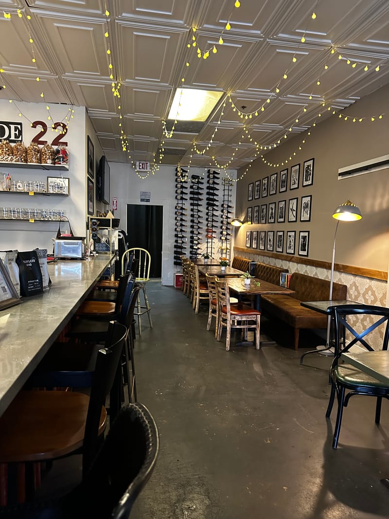 Inside Relentless Grind Coffee: a long counter with stools on one side, tables and a bench along the other, string lights overhead and a wall of wine bottles at the back.