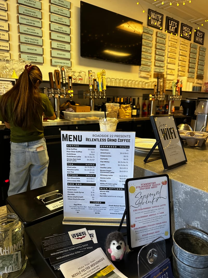 The Relentless Grind Coffee menu standing on the counter inside Roadside 22, a seasonal drinks sign beside it and beer taps behind.